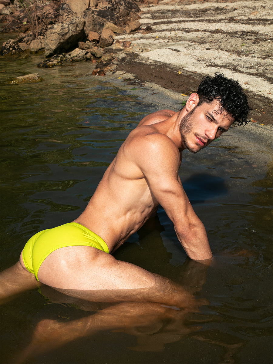 A male model kneeling in shallow water wearing lime green swim briefs.