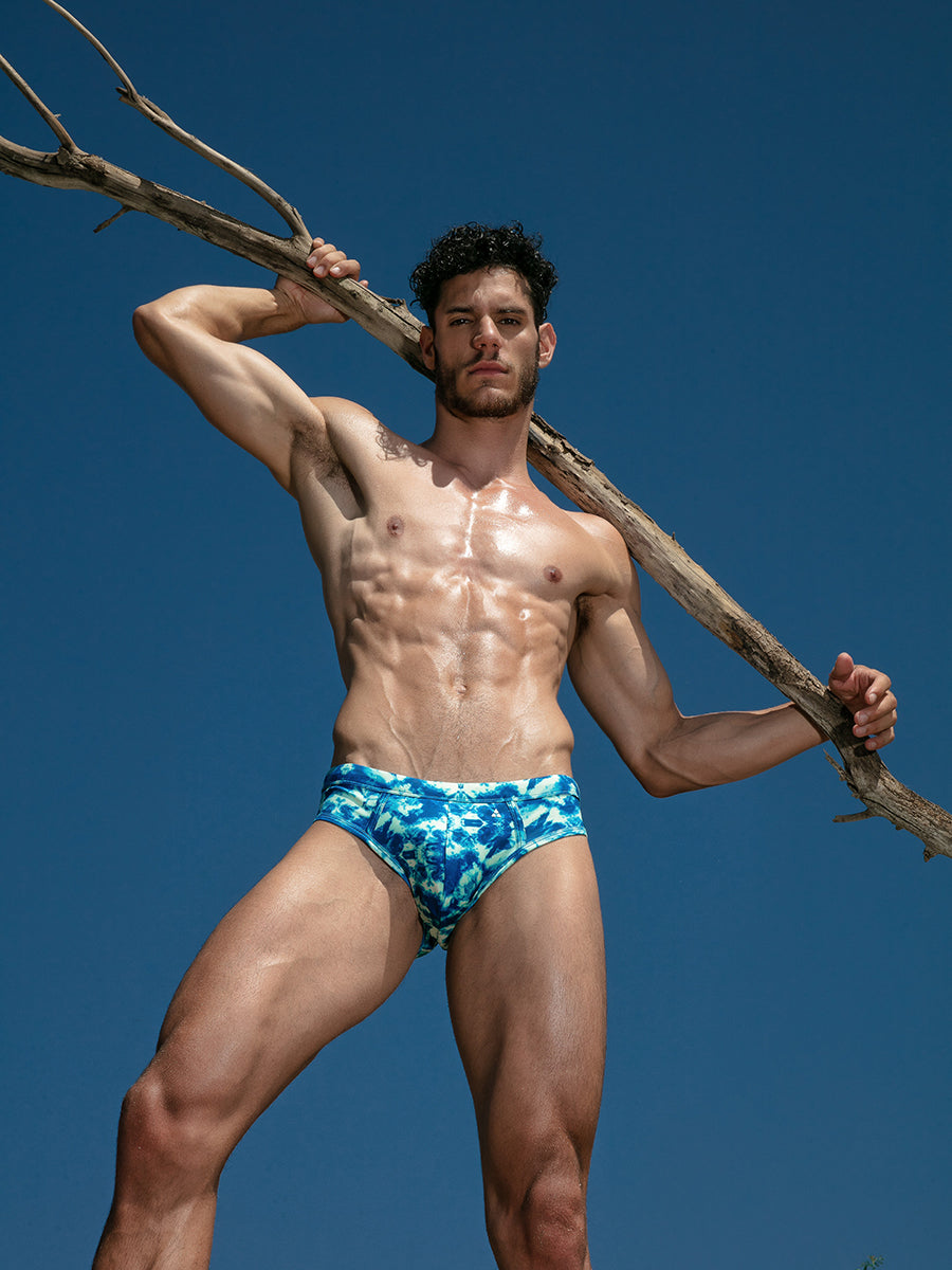 Front view of a male model wearing blue and white tie dye swim briefs, holding a branch behind his head.