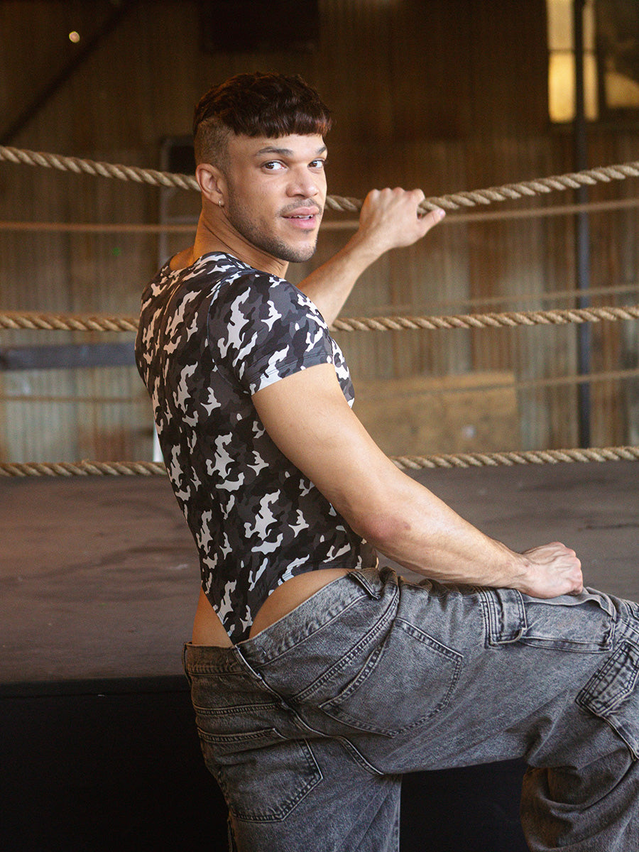 Rear view of a male model wearing a black and white camo thong bodysuit, next to a boxing ring. 