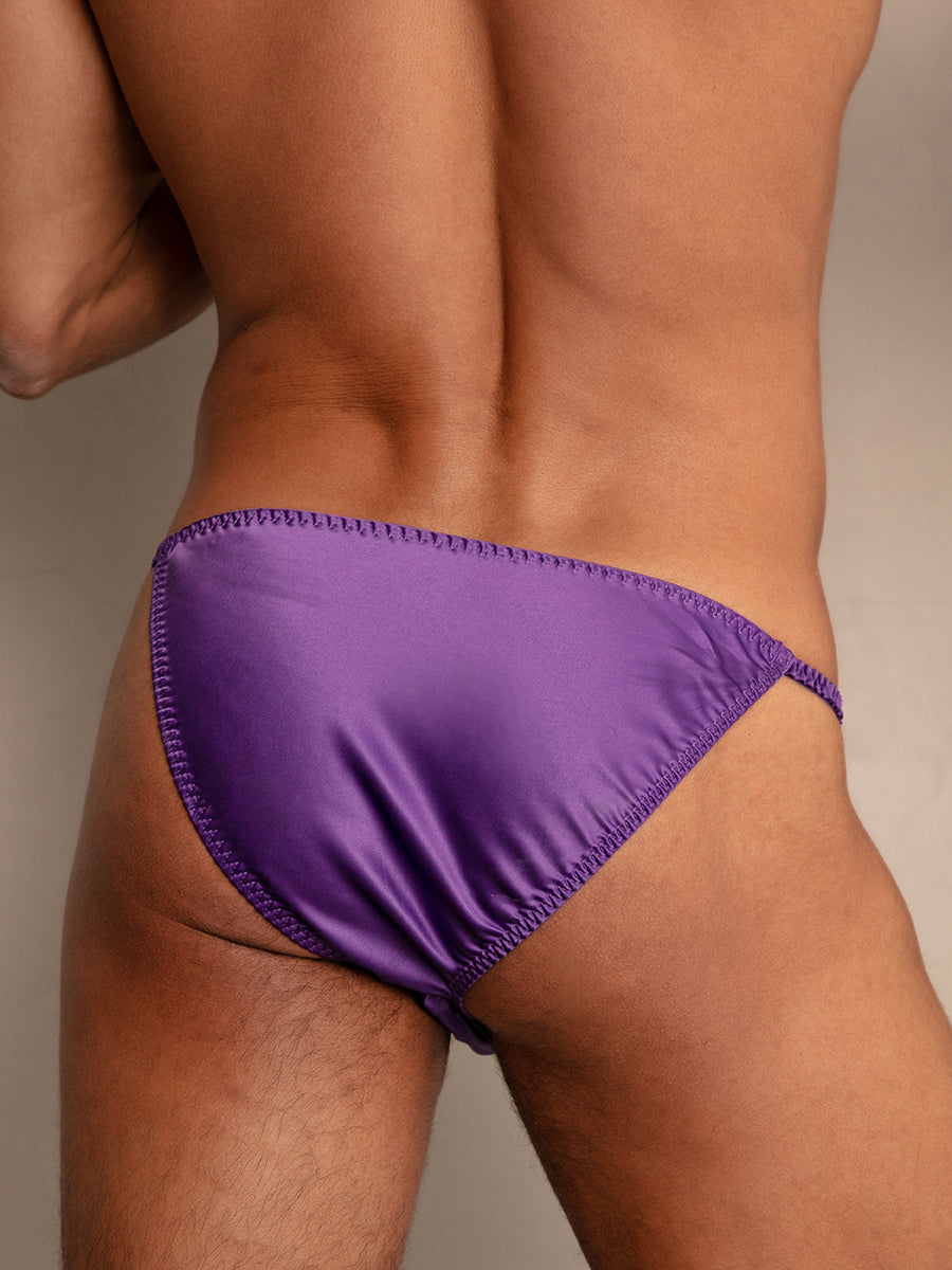 Closeup rear view of a man wearing bright purple bikini style underwear, leaning against a wall.