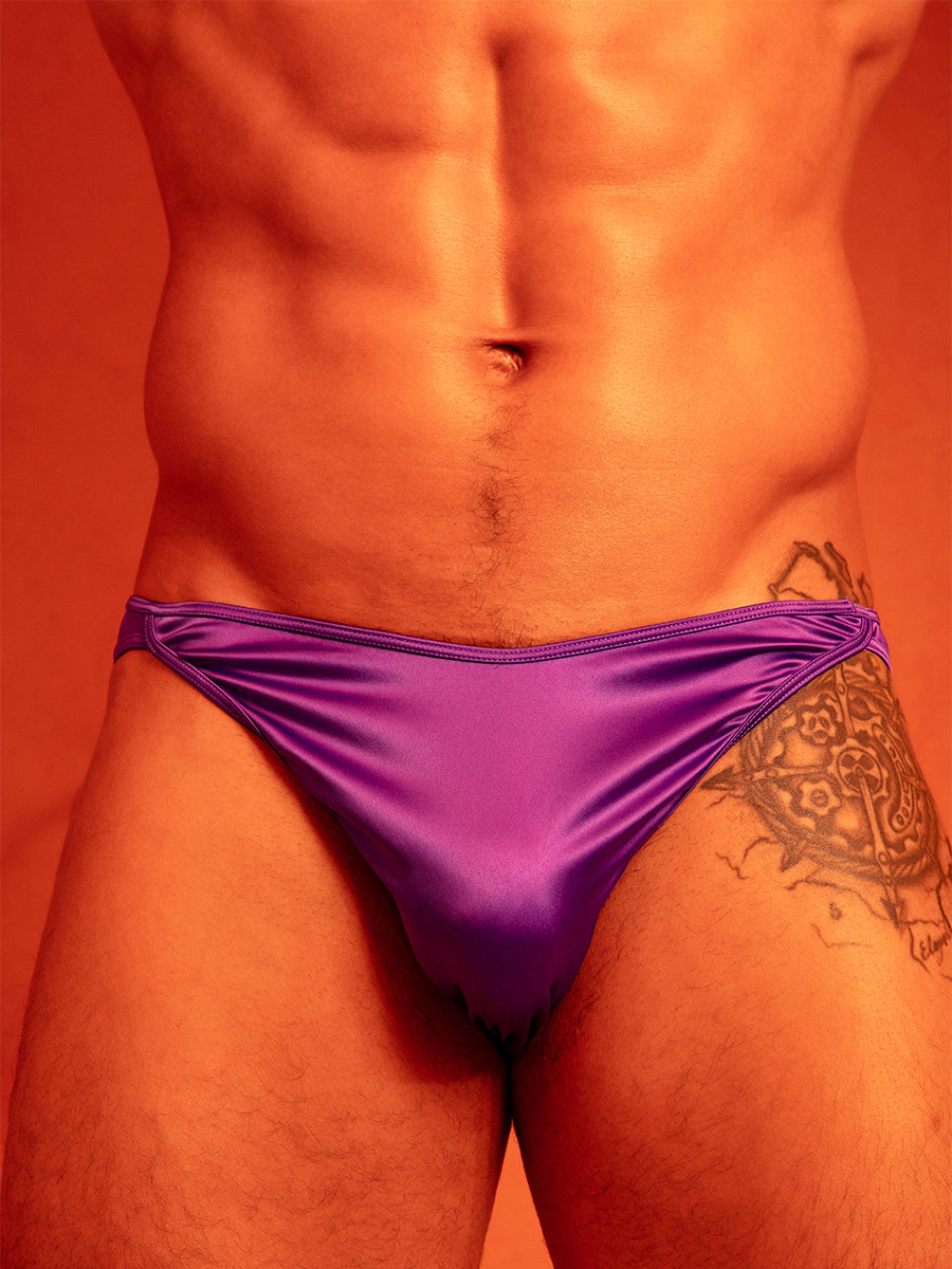 Closeup front view of a man in purple satin underwear briefs, against an orange background.