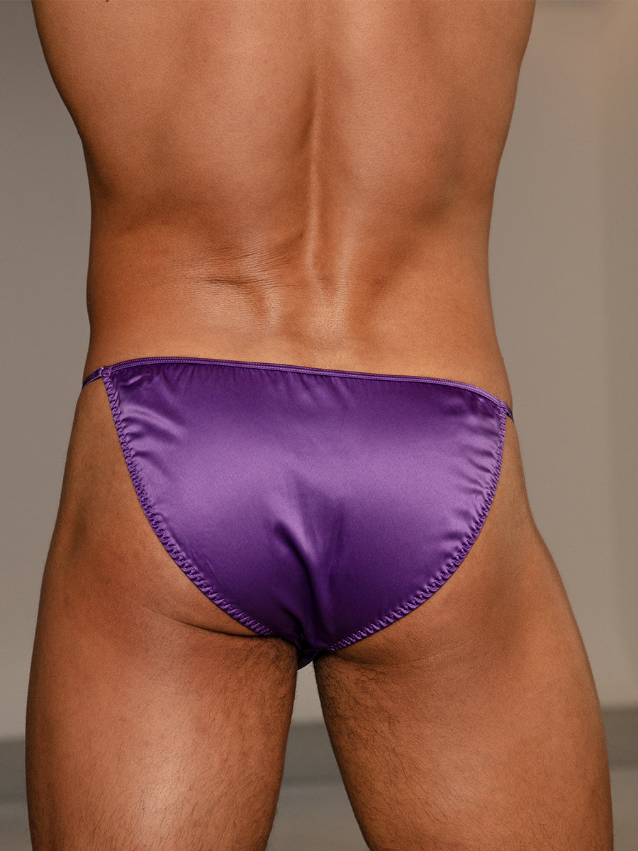 Closeup rear view of a man wearing a purple satin bikini brief against a neutral background.